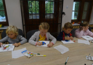 Czworo dzieci siedzi przy stole z mazakami w ręku, kończą malowanie płótna.